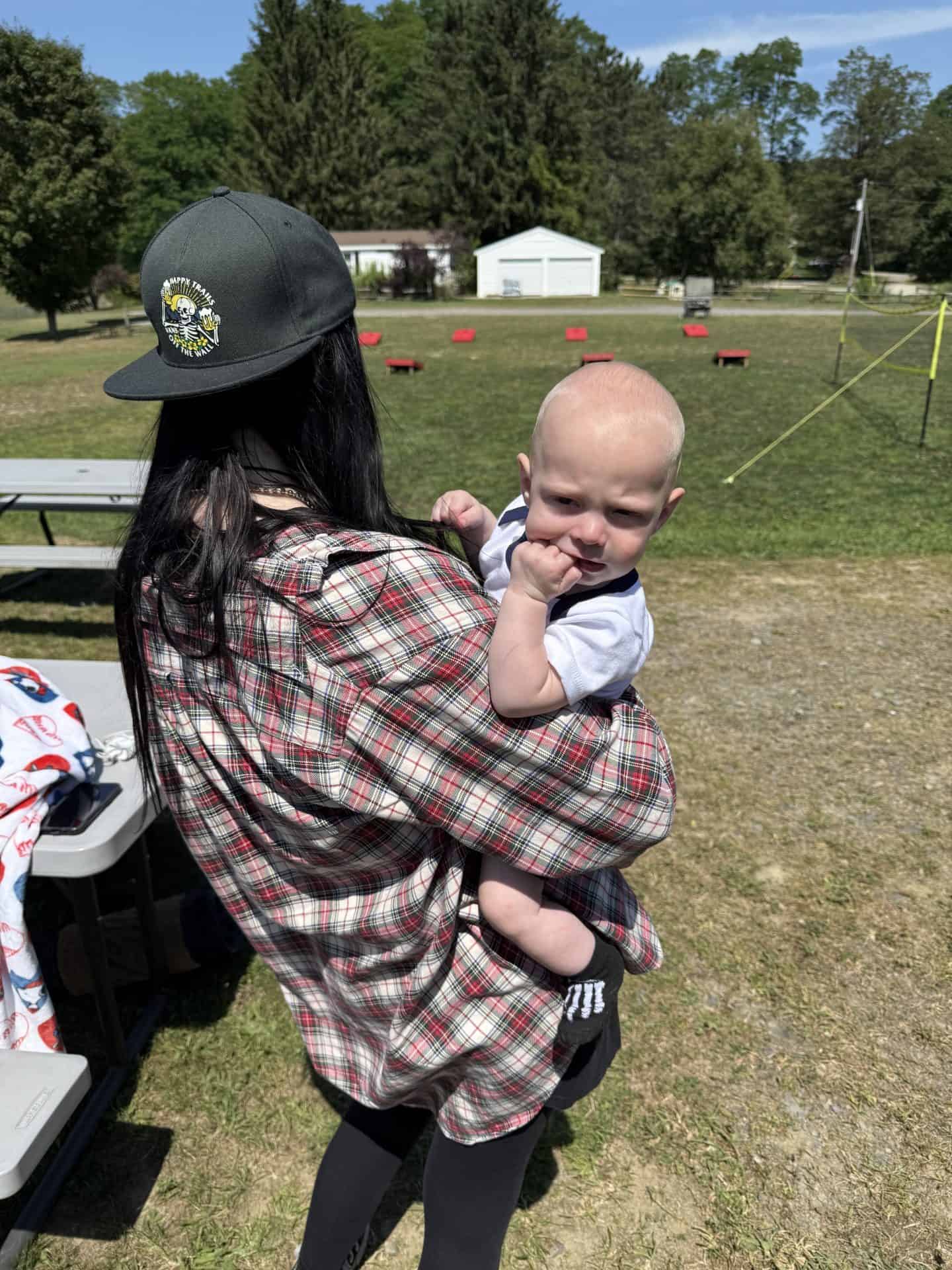 Woman holding a baby outdoors in a park setting with trees and a small building in the background.