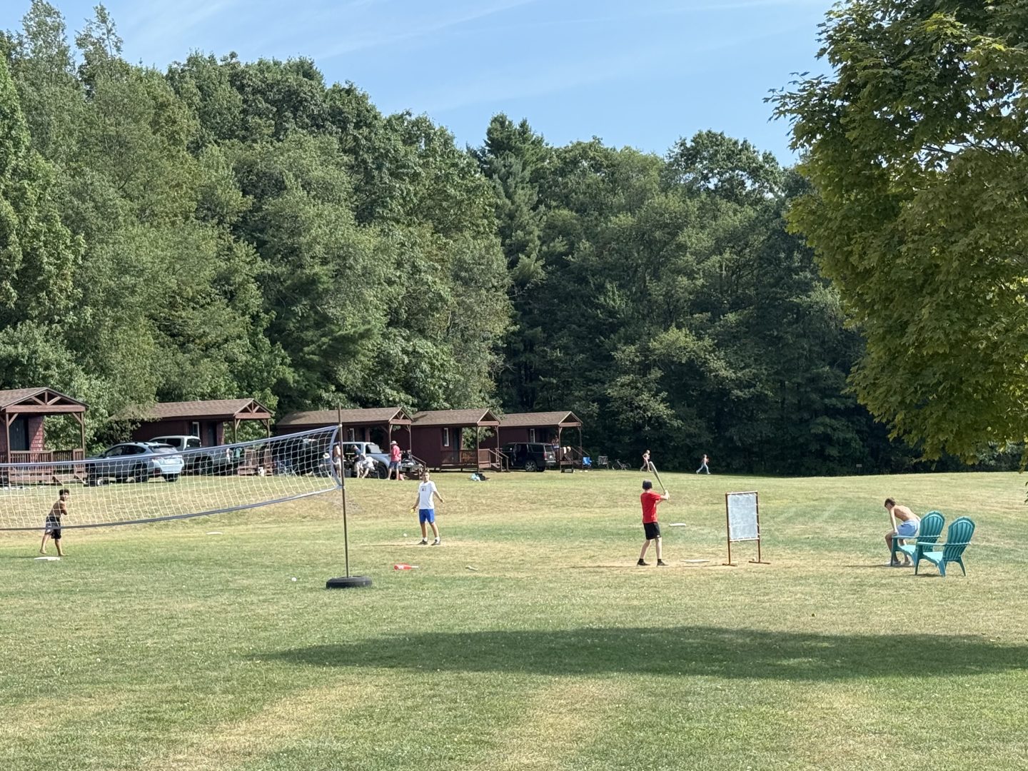 Youth baseball practice on a sunny field with trees and cabins in the background.