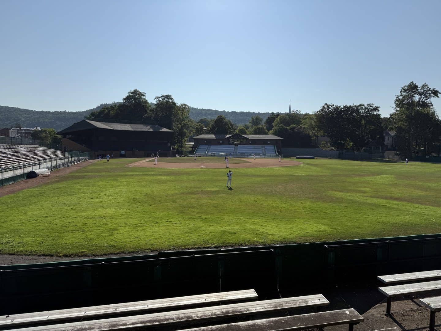Baseball field with players practicing, surrounded by empty stands and scenic trees.