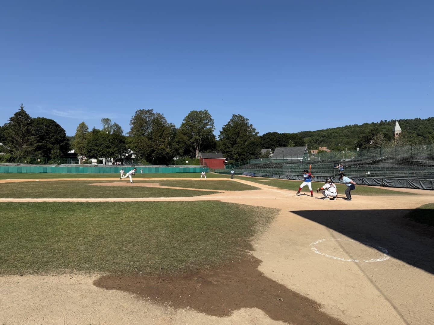 Youth baseball game at a local field with players and umpire under a clear blue sky.