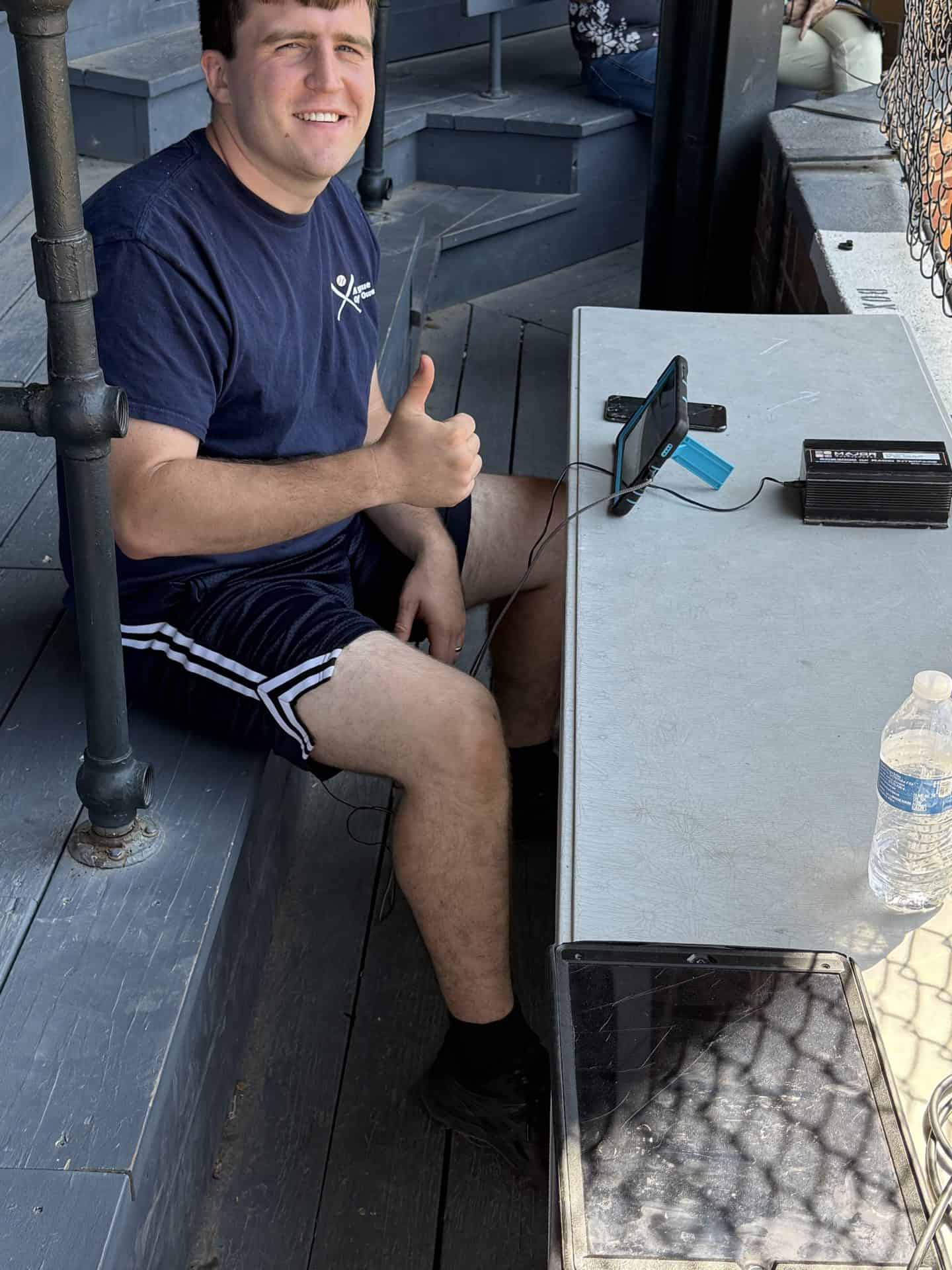 Man sitting outdoors at a table, giving a thumbs-up, with a phone and water bottle nearby.