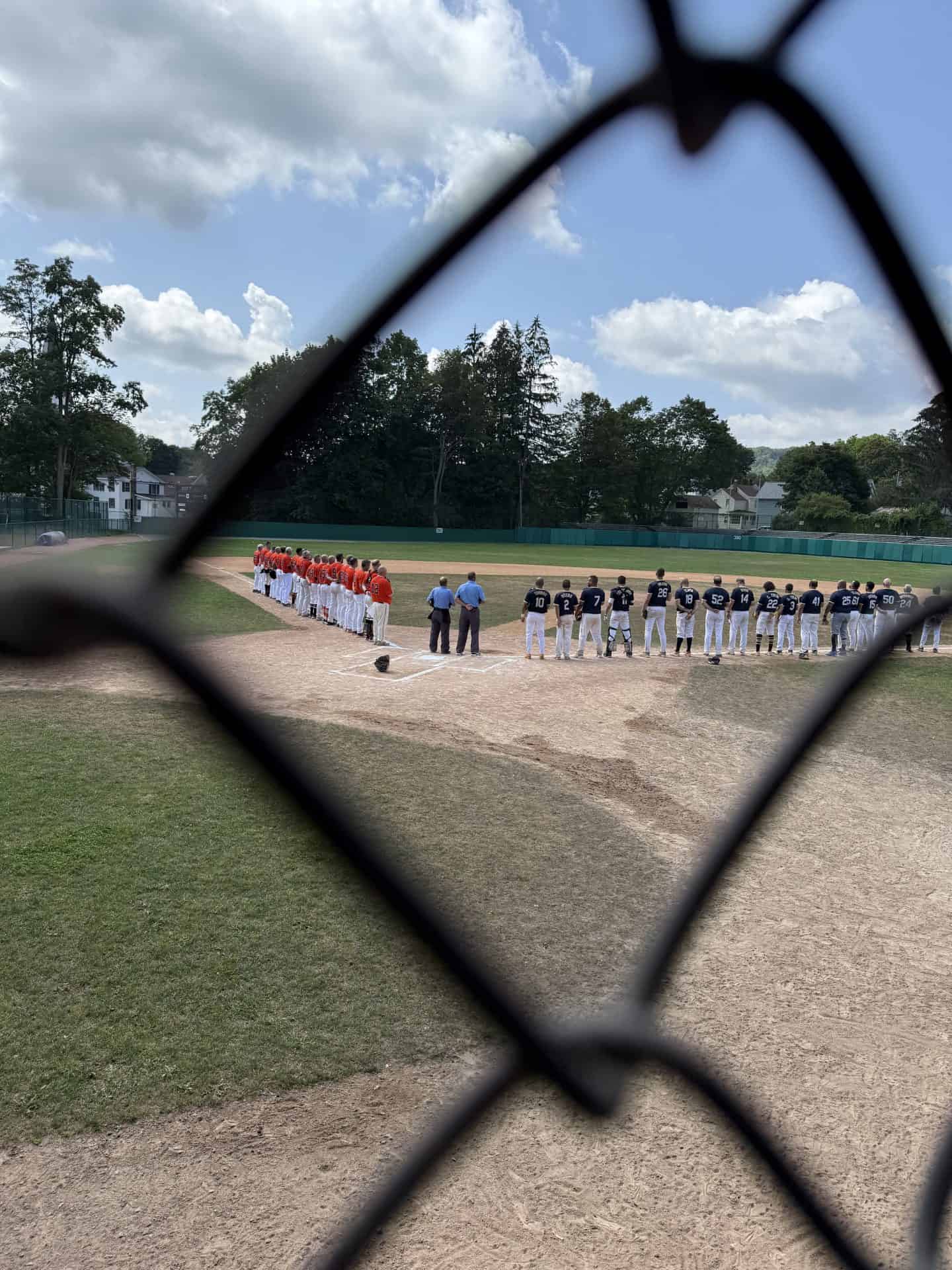 Youth baseball teams lined up on the field before a game, viewed through a chain-link fence.